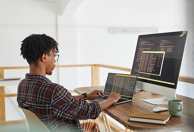 African-American IT developer typing on keyboard with black and orange programming code on computer screen and laptop in contemporary office interior