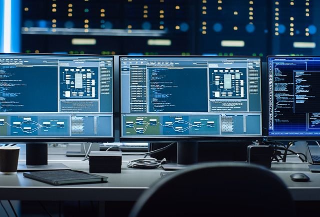 Shot of Multiple Personal Computer Monitors Showing Coding Language Program with System Monitoring Interface. In the Background Data Center with Server Racks.