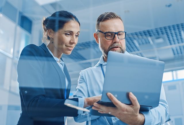 Businesswoman and Businessman Working on a Project, Man Holds Laptop Computer, They Reference it.