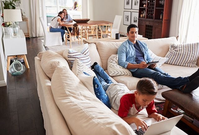 Pre-teen boy lying on sofa using laptop, dad sitting with tablet, mum and grandma in the background